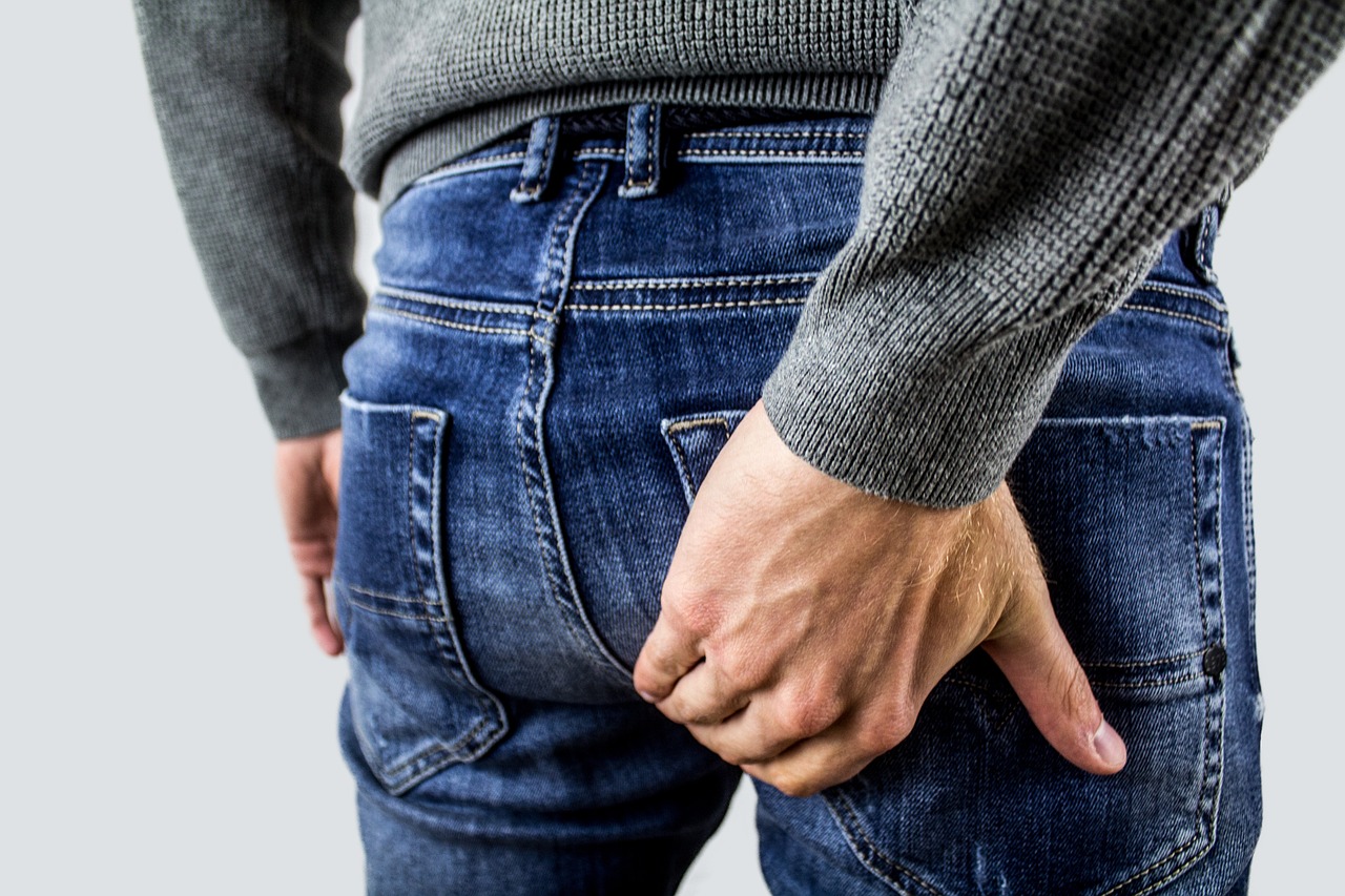 A man in denim jeans holding his lower back, indicating discomfort near his perineum. His gesture of holding his lower bottom suggests he may be dealing with perineal pain, a condition that can significantly impact daily life activities.